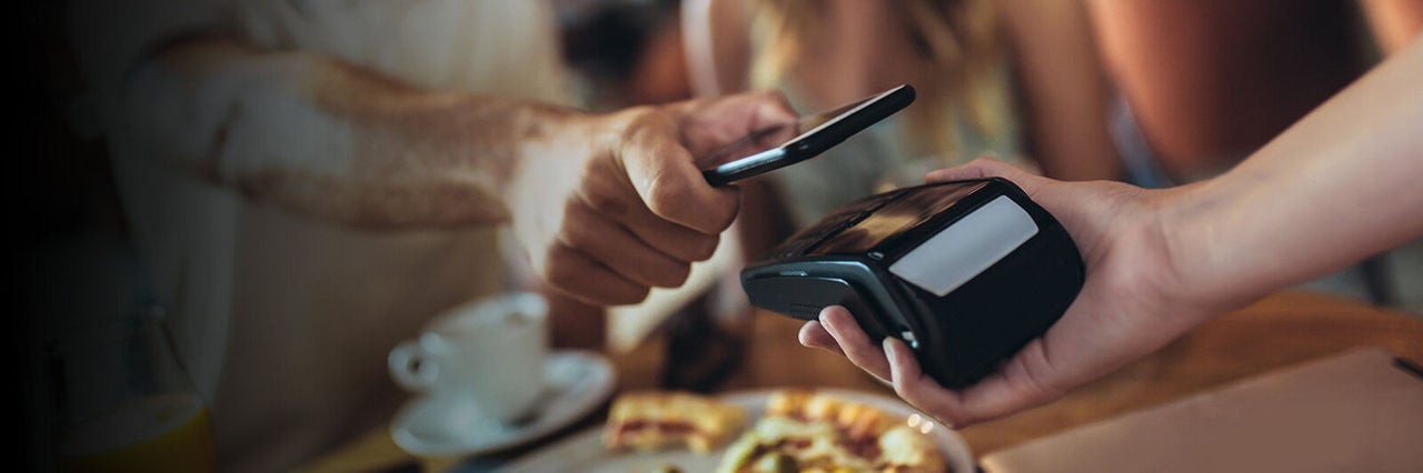 Egy vendég okostelefonnal érintésmentesen fizet egy kézben tartott bankkártya-terminálnál egy étteremben, az asztalon kávé és étel látható.