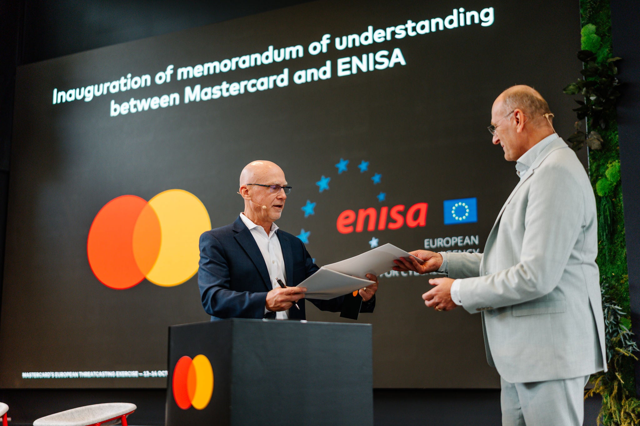 Zwei Männer tauschen während der Unterzeichnung des Mastercard- und ENISA-Memorandums Dokumente auf der Bühne aus.