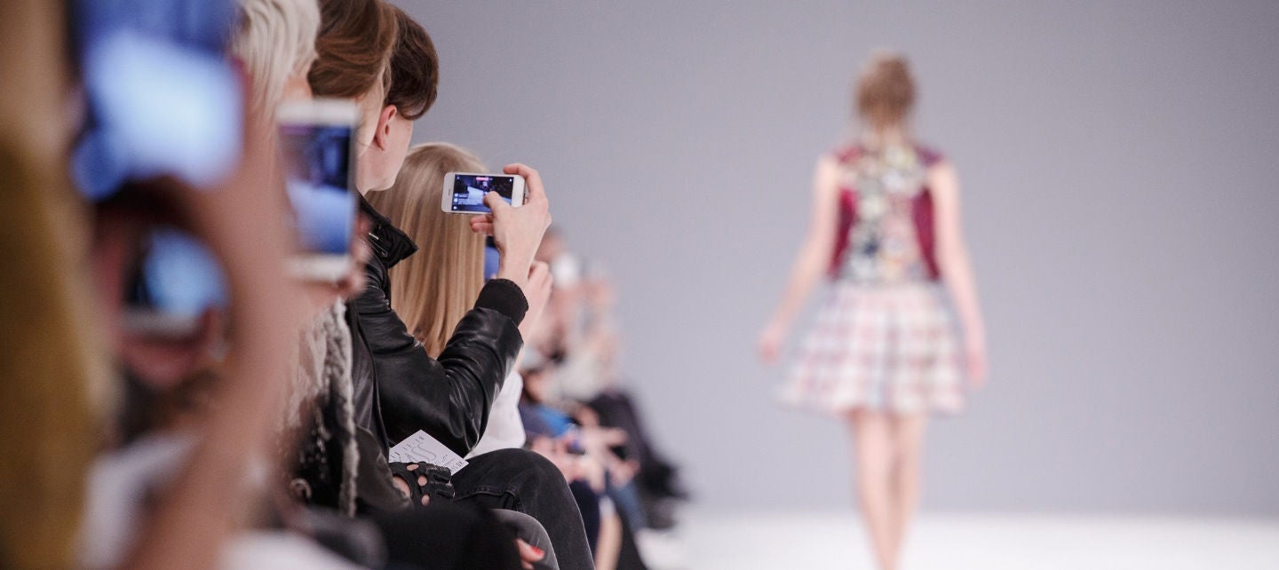 A woman walks down the runway at a fashion show with a row of people holding up cameras. 