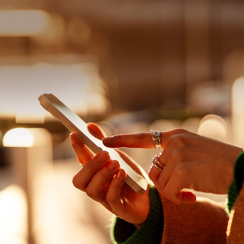 Zbliżenie na dłonie osoby korzystającej ze smartfona na rozmytym, ciepłym tle.