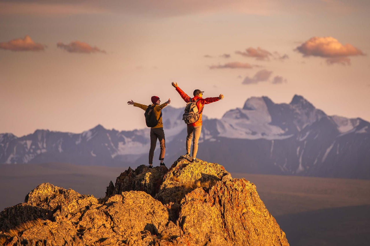 Two hikers stand atop a peak with their arms out overlooking mountains. 