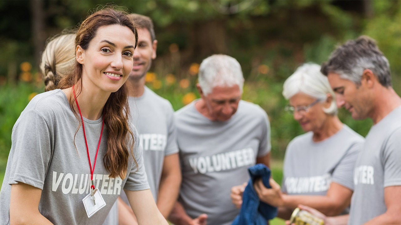 Mosolygó női önkéntes a kép előterében, mögötte különböző korú önkéntesek csoportja szürke „Volunteer” pólóban, szabadtéri közösségi programon, zöld környezetben.