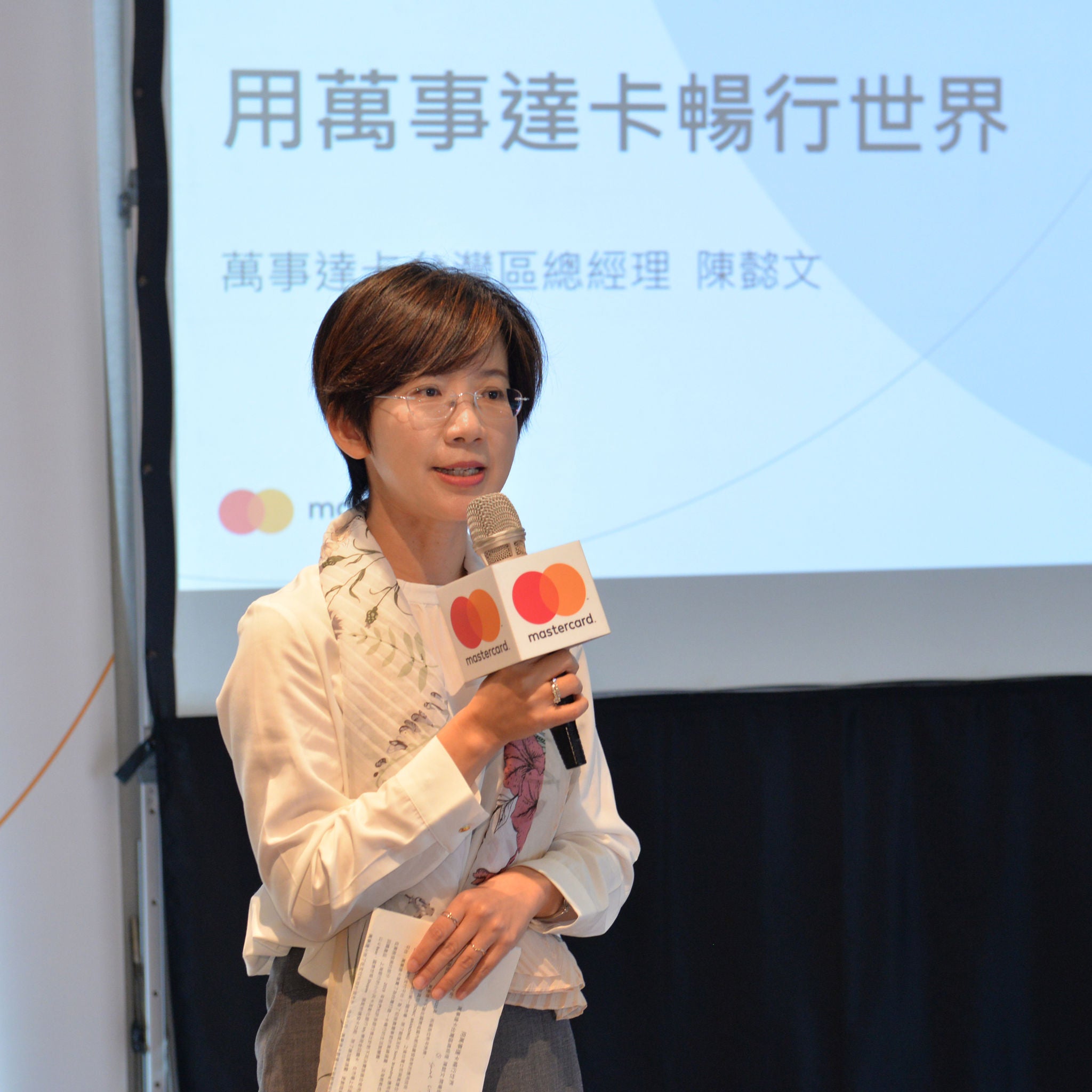 A woman in a white blouse speaks into a handheld microphone on stage in front of a projection screen with Chinese text.