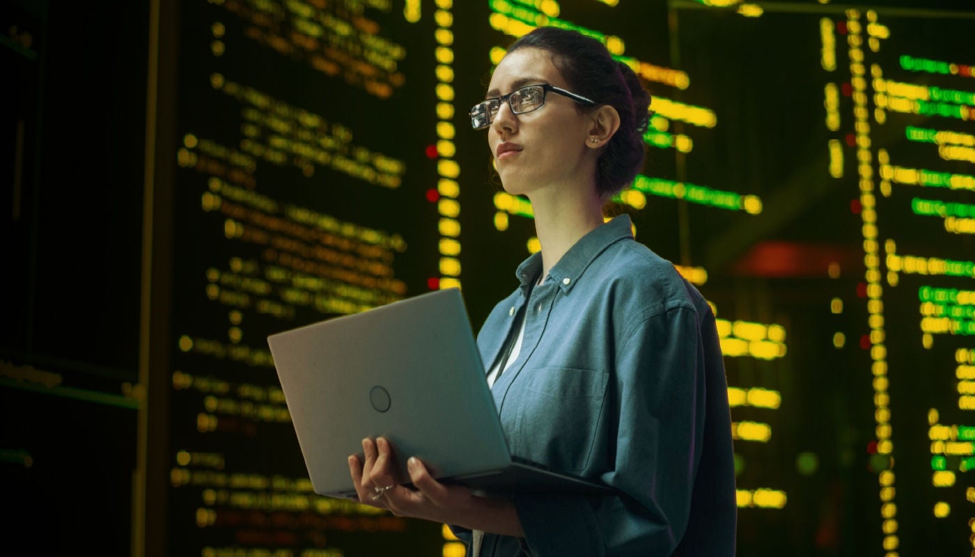 A woman reviewing code on big screens around her while on her laptop