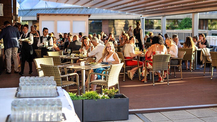 Vendégek ülnek asztaloknál egy fedett teraszon, italokkal és ételekkel, személyzet szolgál fel a háttérben