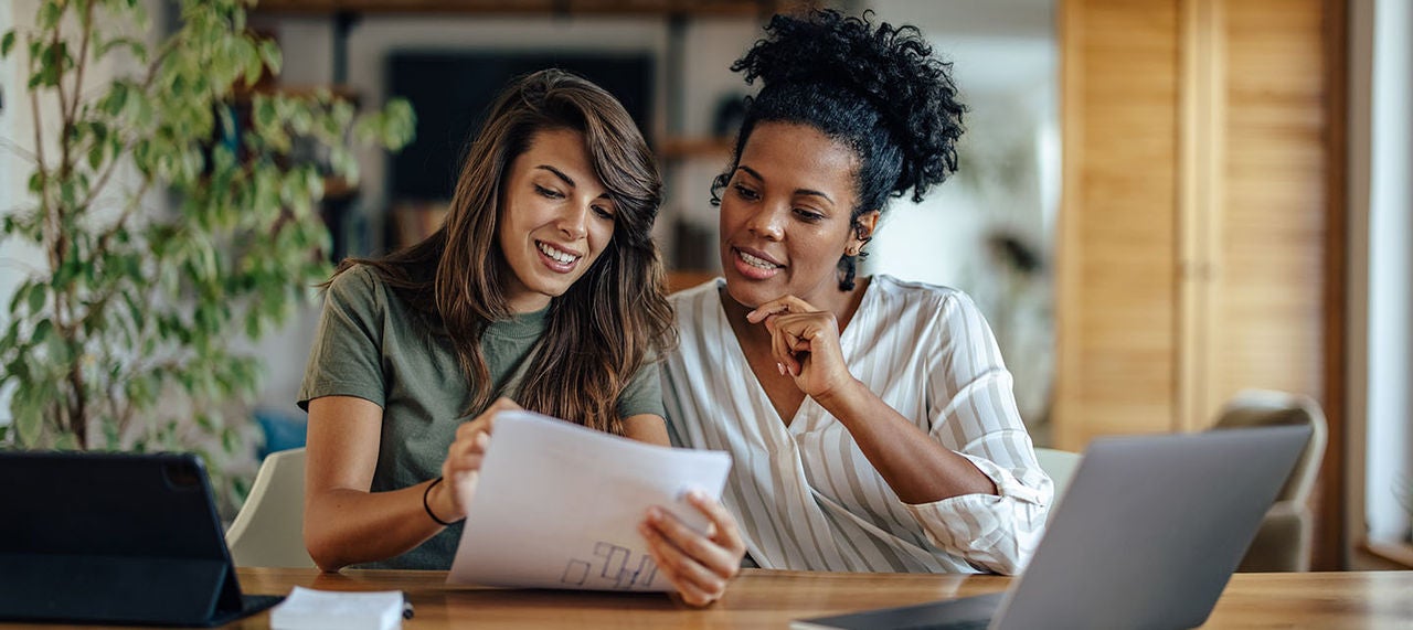 Two women sitting at a table with a laptop and documents, collaborating on financial planning