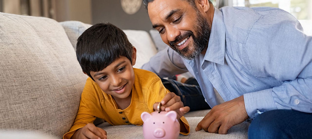 Father helping young son save money by placing coins into pink piggy bank at home.