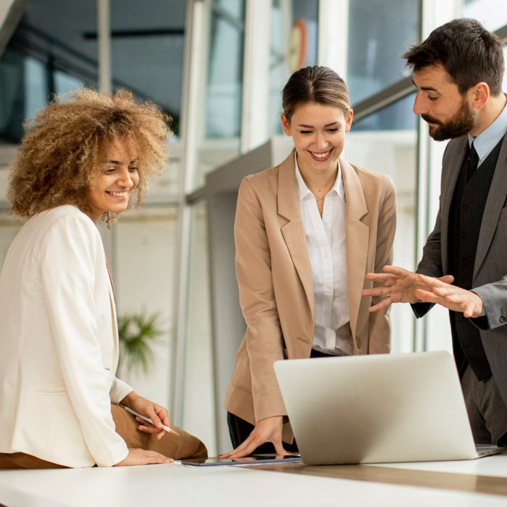Three smiling business professionals, two women and one man, collaborating around a laptop in a modern office.