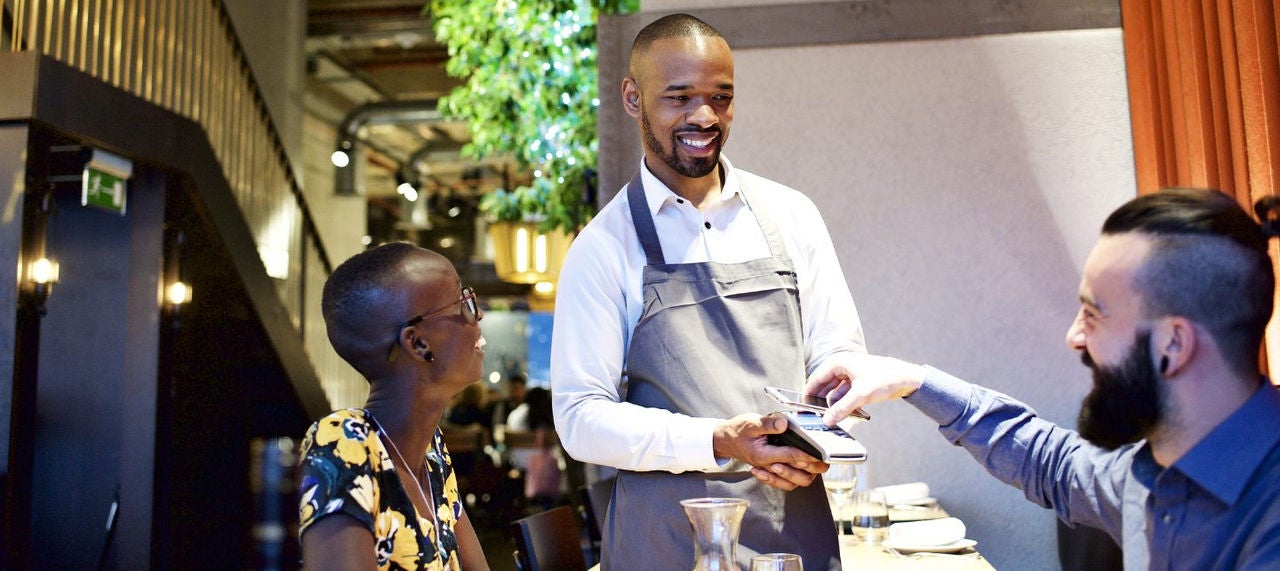 A man holds his mobile wallet to a POS machine held by a waiter in a restaurant. 
