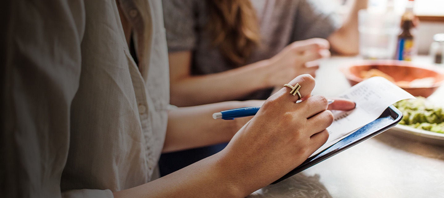Nærbilde av hender som skriver på papir med penn, andre personer i bakgrunnen.