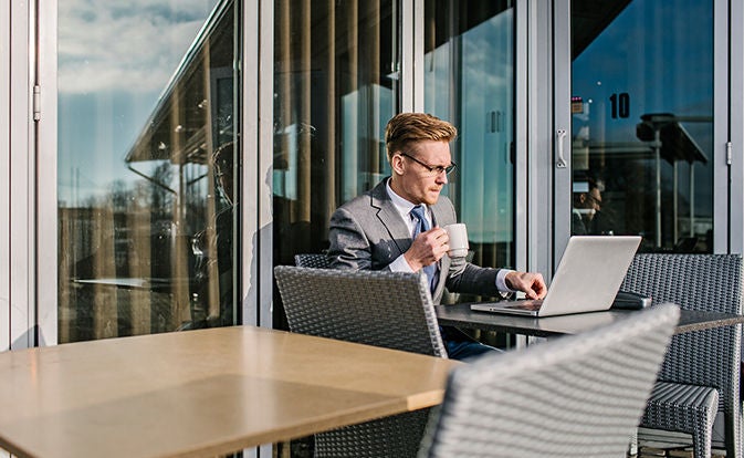 Man in a suit sitting outdoors with a coffee, working on a laptop.