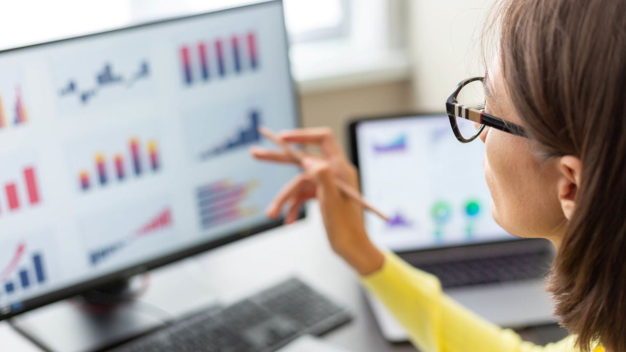 Woman with spectacles and yellow cardigan holding a pen and looking at a monitor with various graphs displayed.