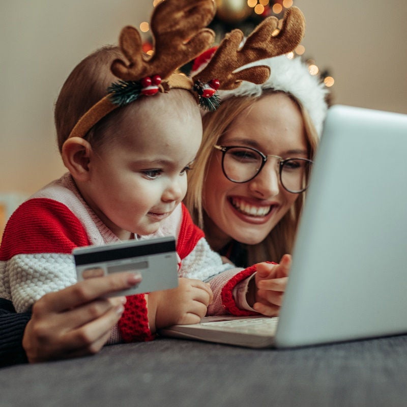 A woman in a Santa hat and a young girl in an antler headband look at a laptop while the woman holds a credit card. 