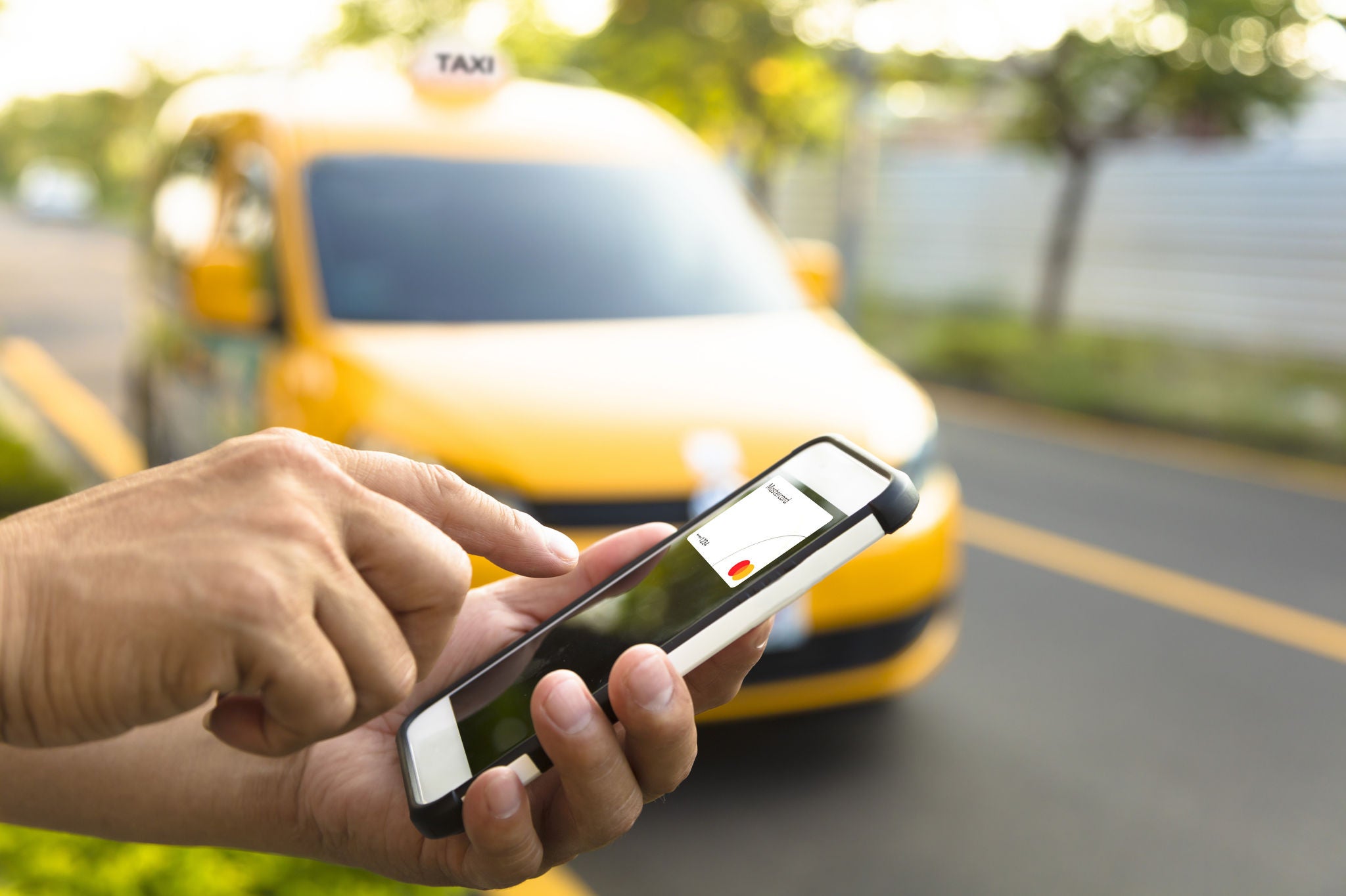 A close-up of hands holding a phone displaying a payment card on screen, with a yellow taxi blurred in the background.