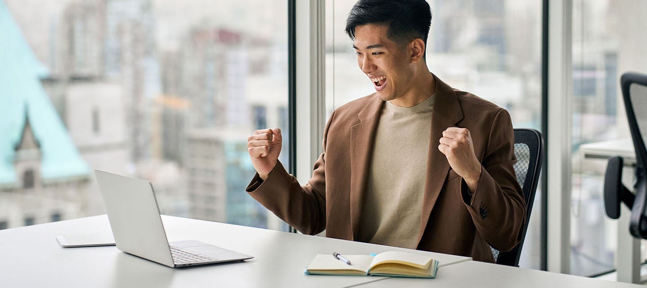Excited young man celebrating success in front of laptop at office desk with city view background.