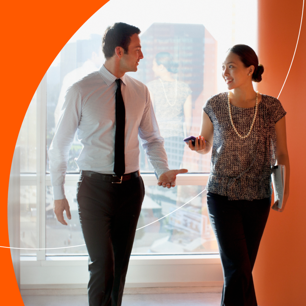 Two business professionals, a man and a woman, walking and conversing in a modern office.