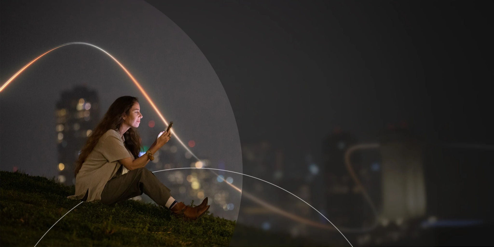 אישה יושבת על דשא בלילה, משתמשת בטלפון שלה, כשברקע אורות עיר מטושטשים זוהרים.
