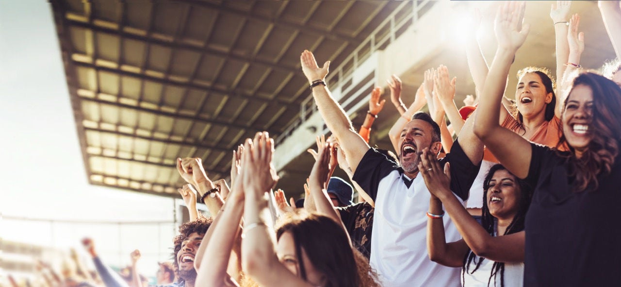 Crowd of sports fans cheering during a match in stadium. Excited people standing with their arms raised, clapping, and yelling to encourage their team to win.