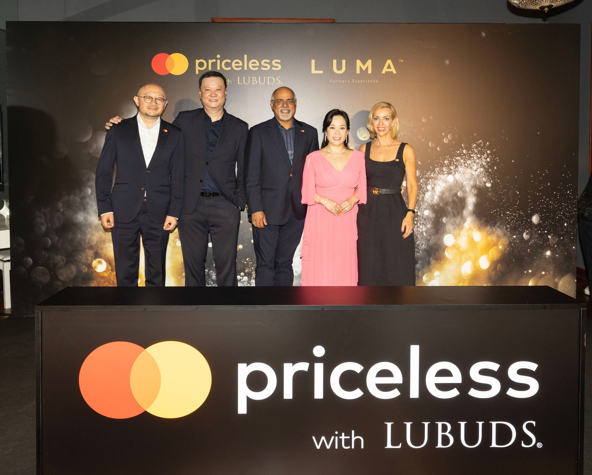 Five people in formal attire stand together in front of a dark event backdrop with brand logos, behind a lit podium.