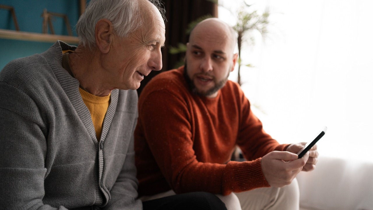 A younger man holds a phone and talks to an older man on a couch. 