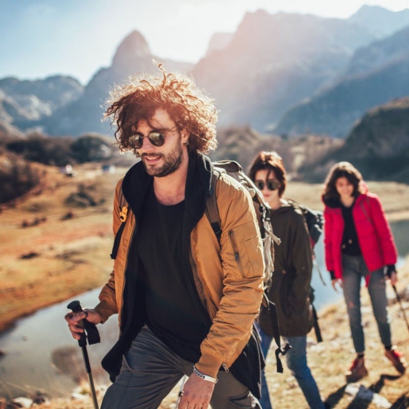 A group of young people in hiking gear and sticks walk along rugged terrain next to a river. 