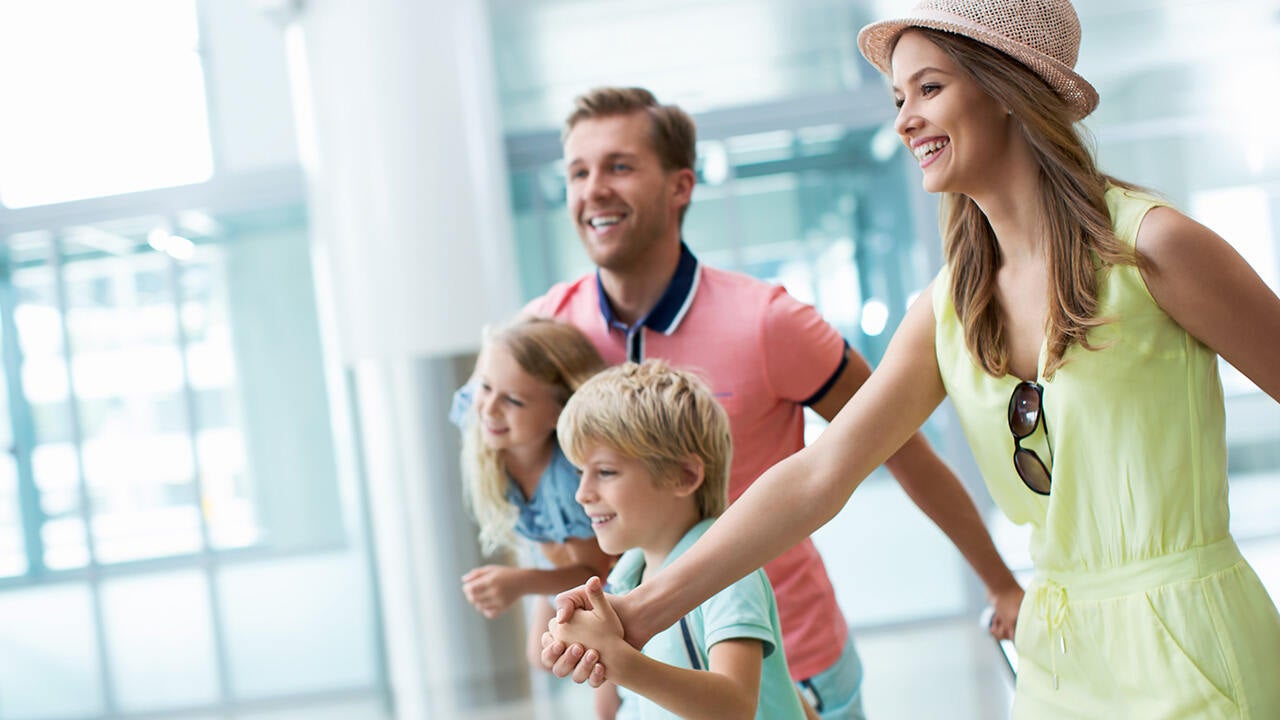 Mosolygó család két gyermekkel világos beltéri térben, közös élményre indulva, utazás vagy szabadidős program hangulatában