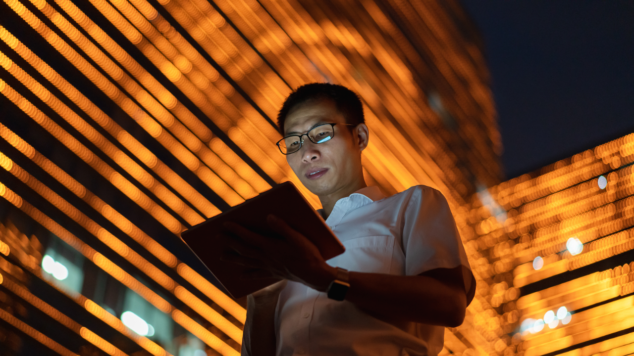 A man looks down at a tablet in front of a brightly-lit building at night. 