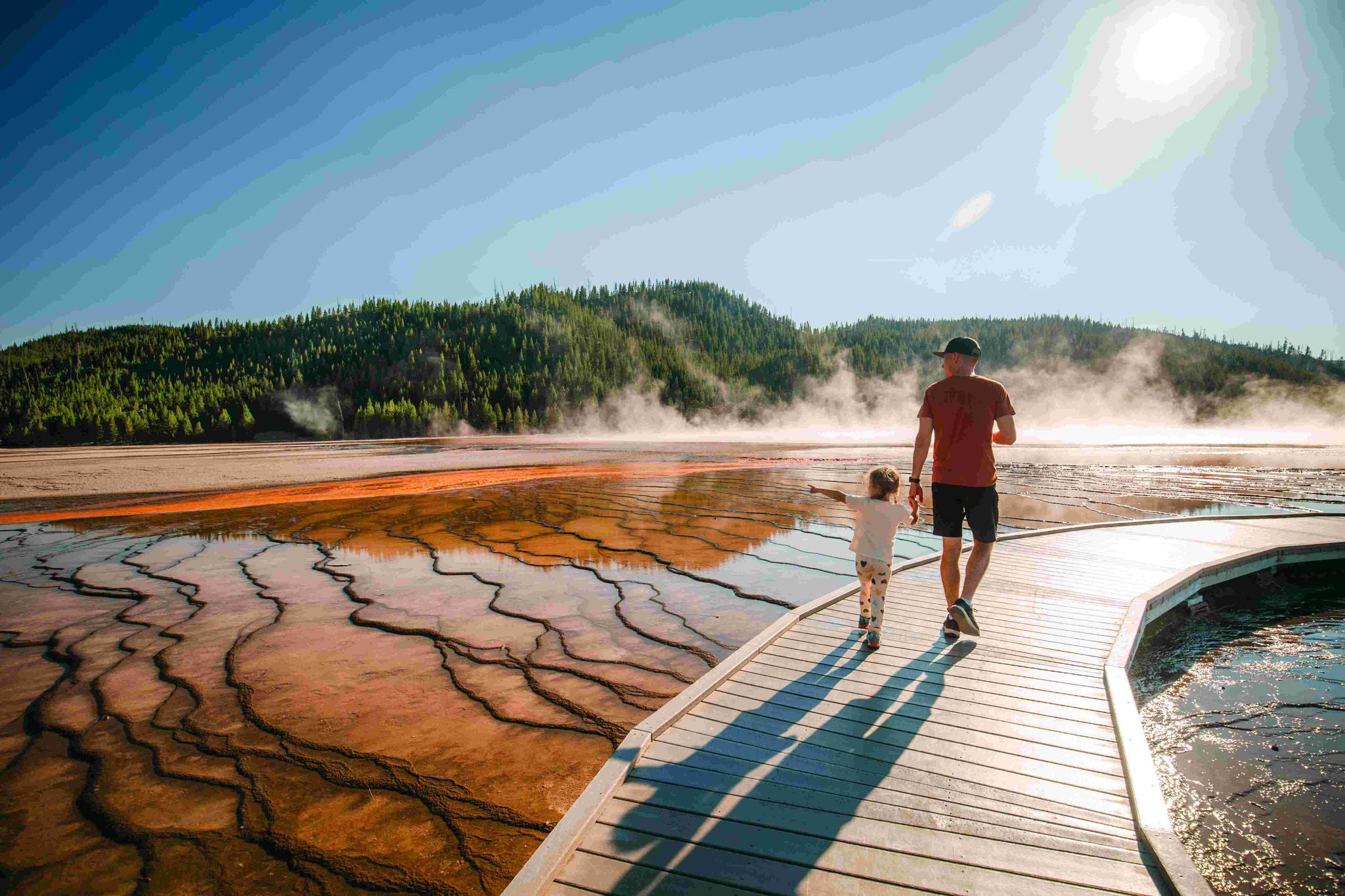 A man and a young girl walk through Yellowstone National Park with steam rising from geysers in the distance. 