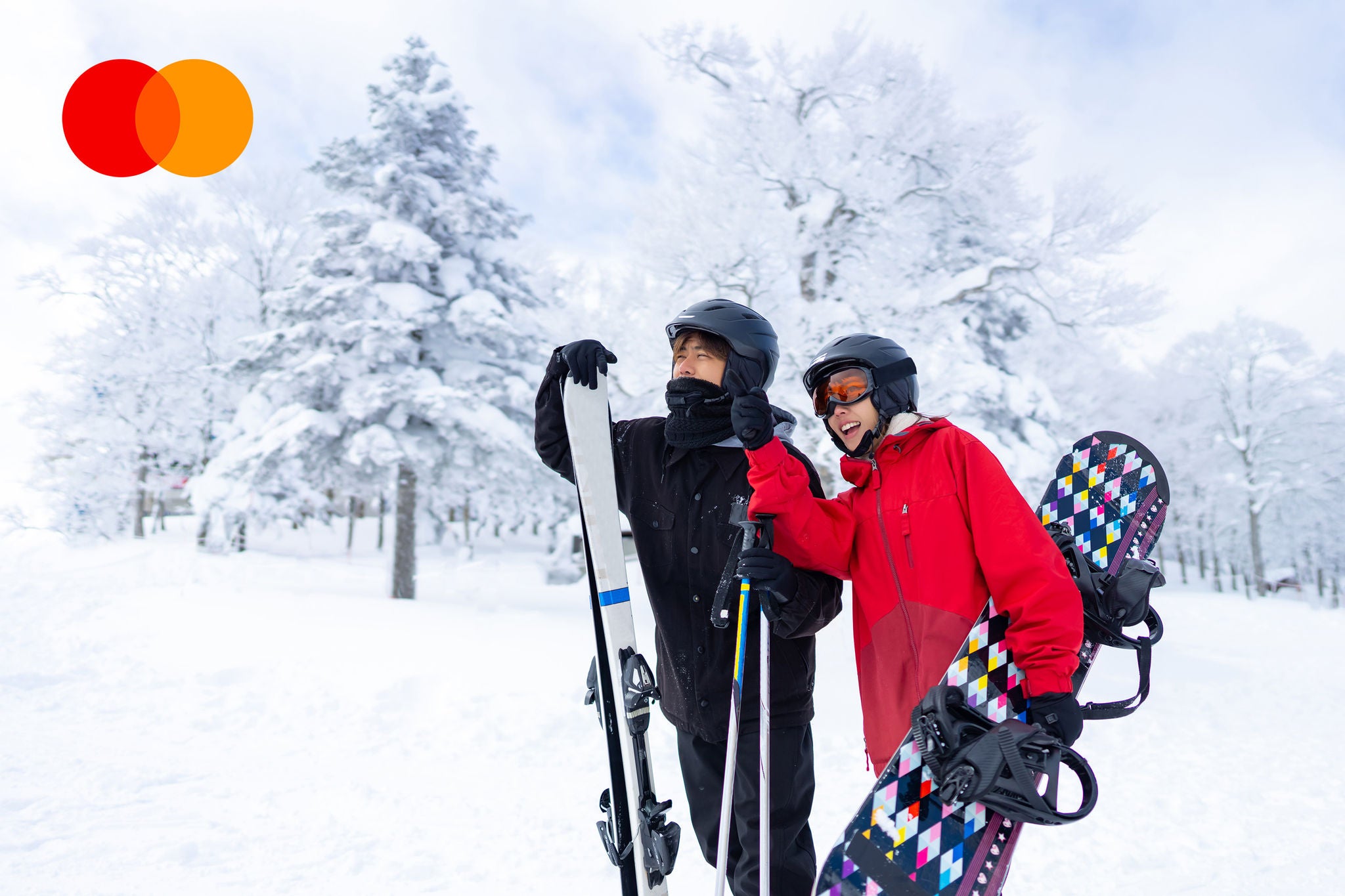 A man holding skis and a woman holding a snowboard stand together on a snowy slope surrounded by snow-covered trees.