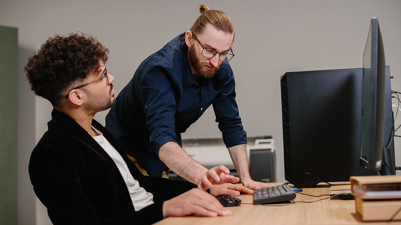 Two male colleagues adjusting settings on desktop computer.