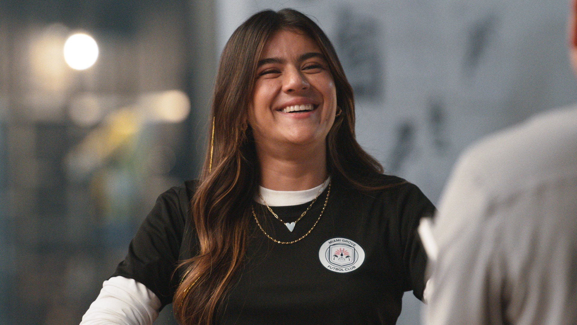 Sofia Carvajal smiles widely at the camera. She wears a black t-shirt with a "Miami Grove Futbol Club" logo over a white long-sleeved shirt, and layered necklaces with a light blue heart pendant.