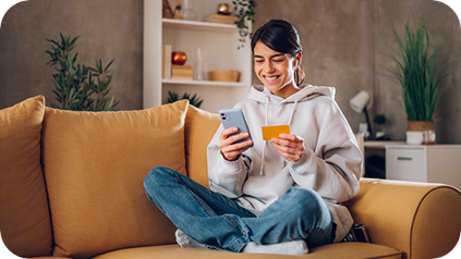 A woman sitting on a couch, holding her phone and a credit card, appears focused on making a purchase.