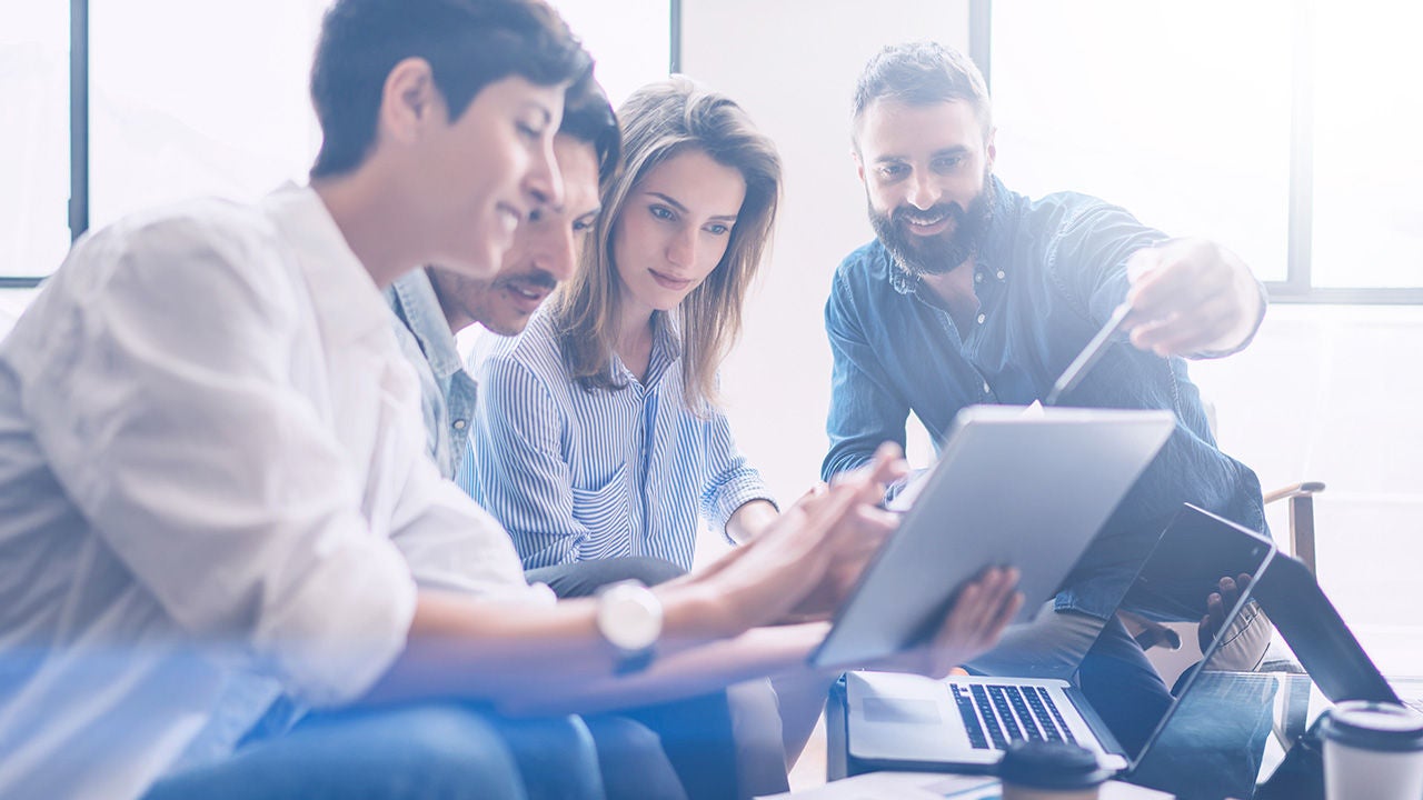 Négy munkatárs világos irodában, közösen egy tabletet és laptopot nézve egy csapatmegbeszélés során.