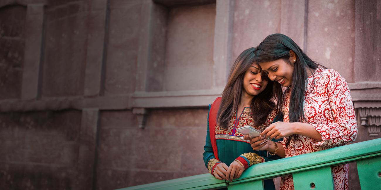 Két fiatal nő hagyományos mintás ruhában egy zöld korlátnál állva mosolyogva néz egy okostelefont, történelmi épület kőfala előtt.