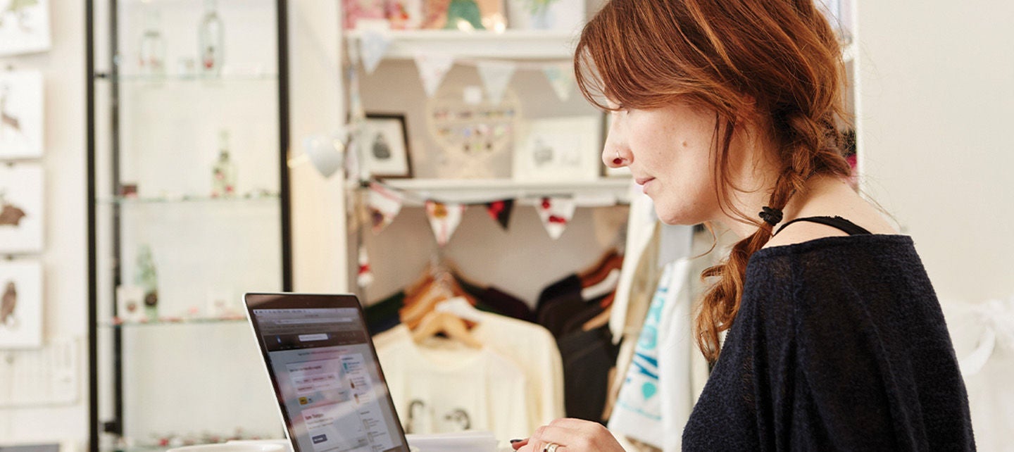 Person med langt hår jobber på en bærbar datamaskin ved et bord i et studio eller butikk, omgitt av kunstverk, klær og dekorasjoner. På bordet står en kopp og noen papirer.