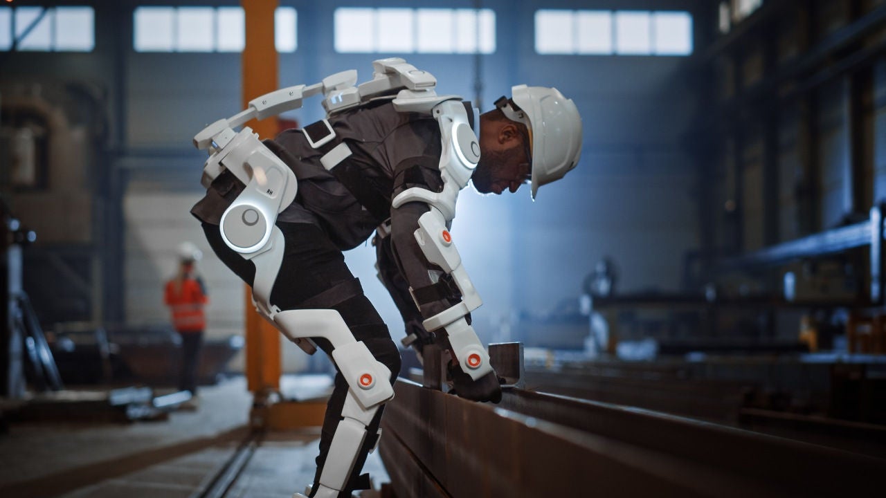 A man wearing an exoskeleten bends over to lift something in  an industrial building site.