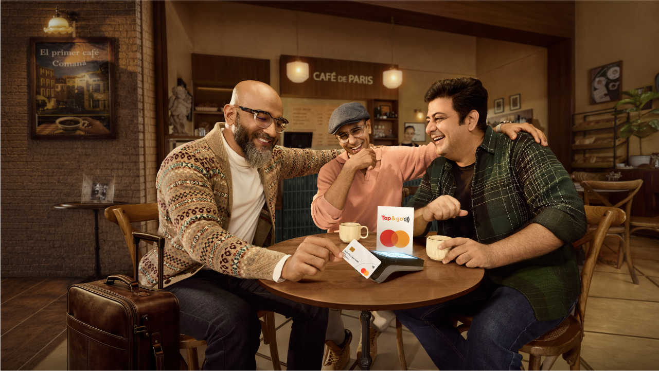 Three indian friends enjoying at a café and one friend making a payment by tapping his Mastercard on the POS machine on their table.