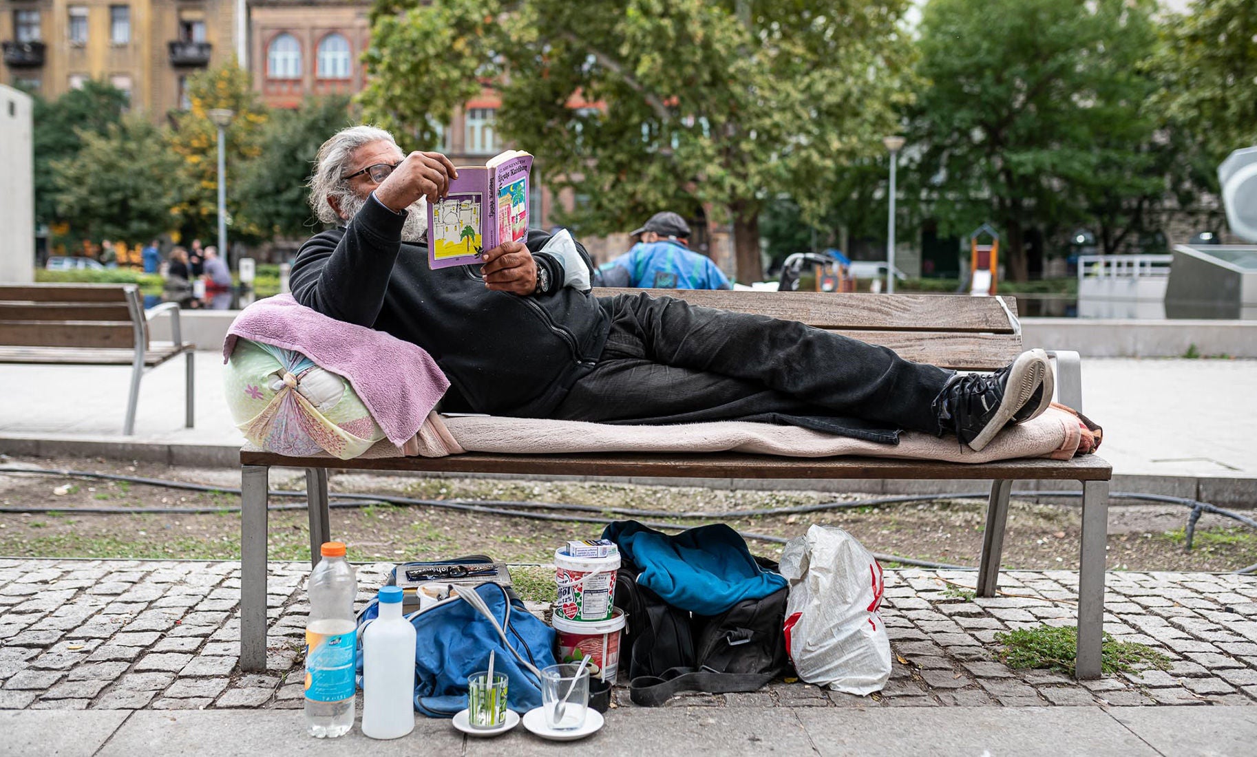 Egy idős férfi egy parkban padon fekve olvas könyvet, a pad alatt személyes tárgyak és palackok vannak elhelyezve.