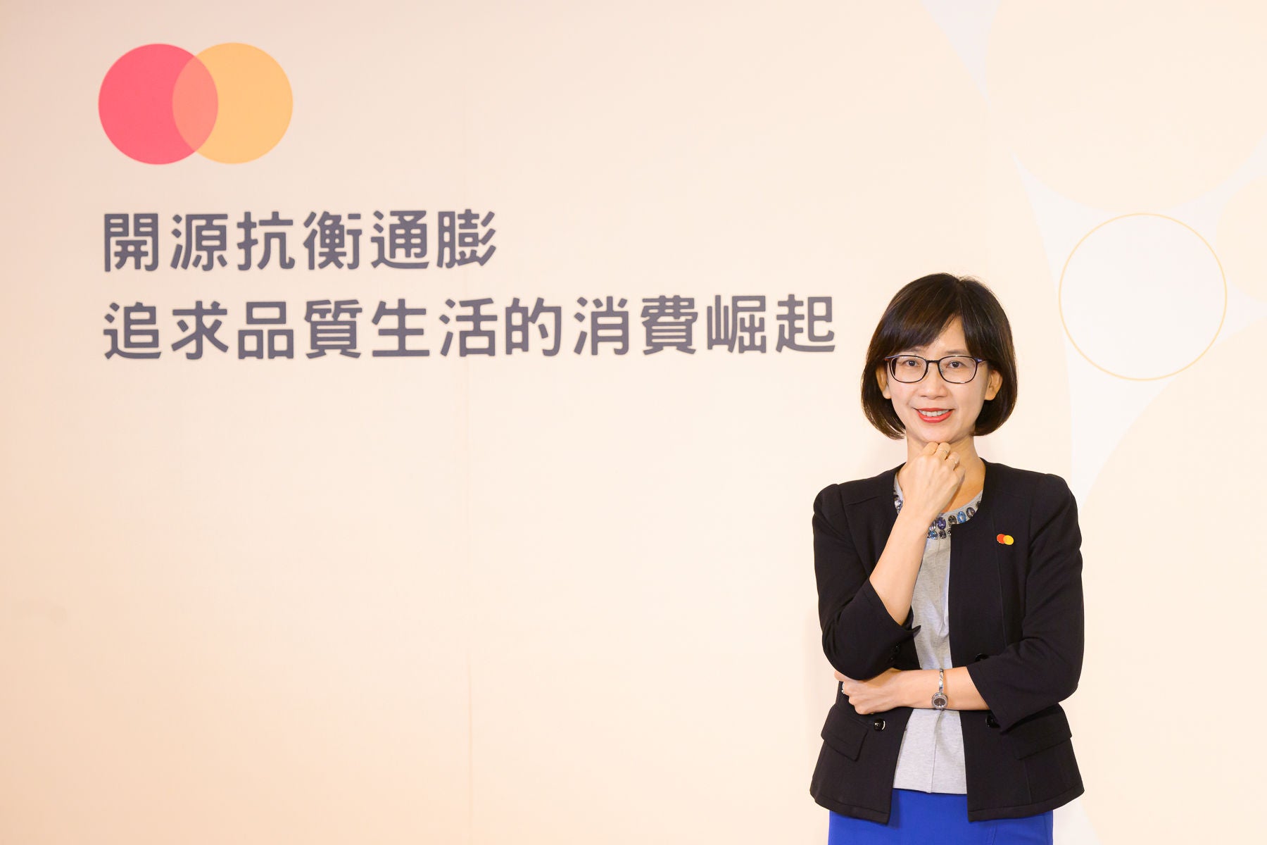 A woman in a black blazer stands with one hand raised to her chin, smiling in front of a cream backdrop with Chinese text and a brand logo.