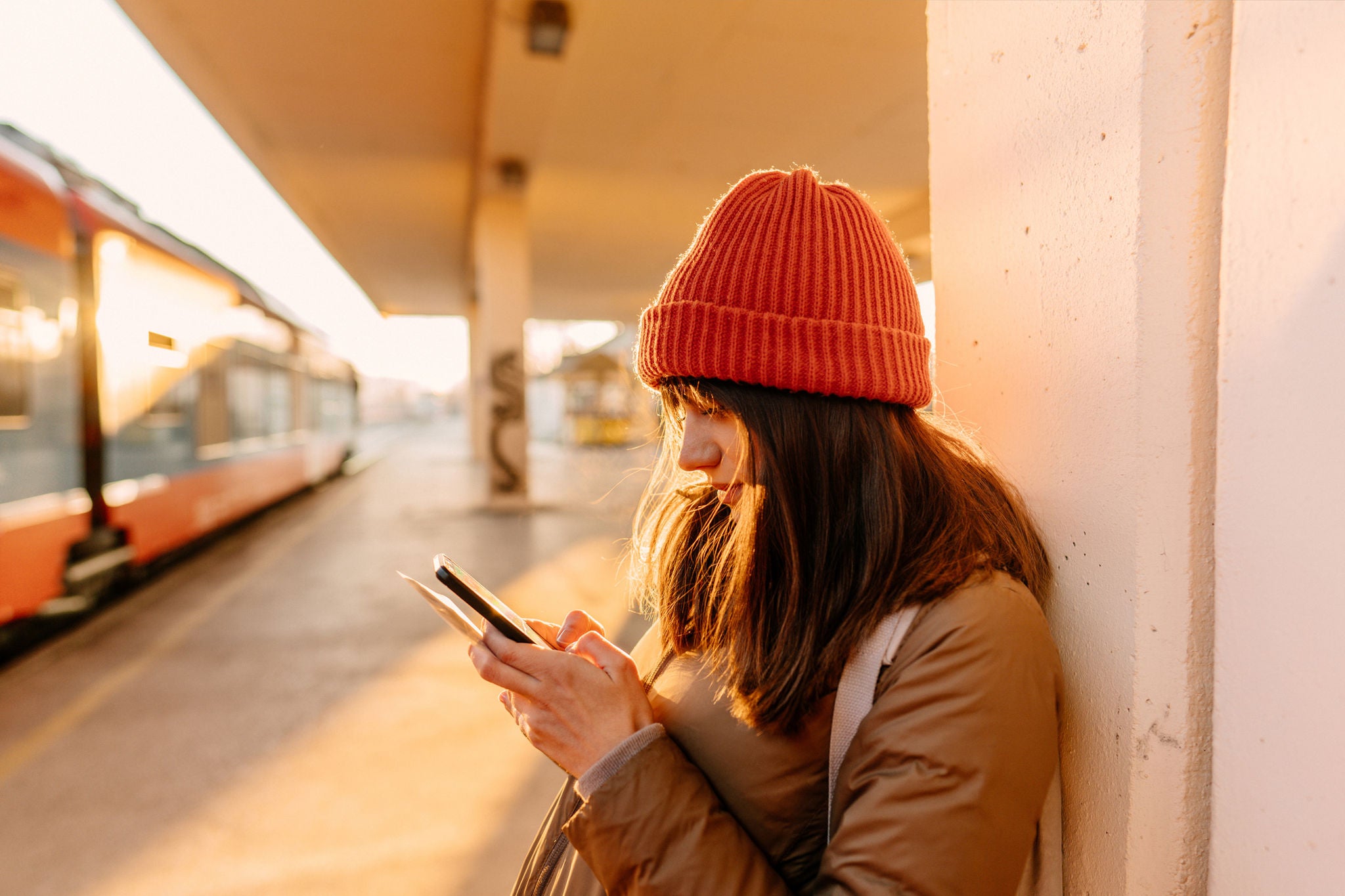Side view of a young woman wearing an orange beanie and jacket, standing on a sunny train platform while looking at her smartphone.