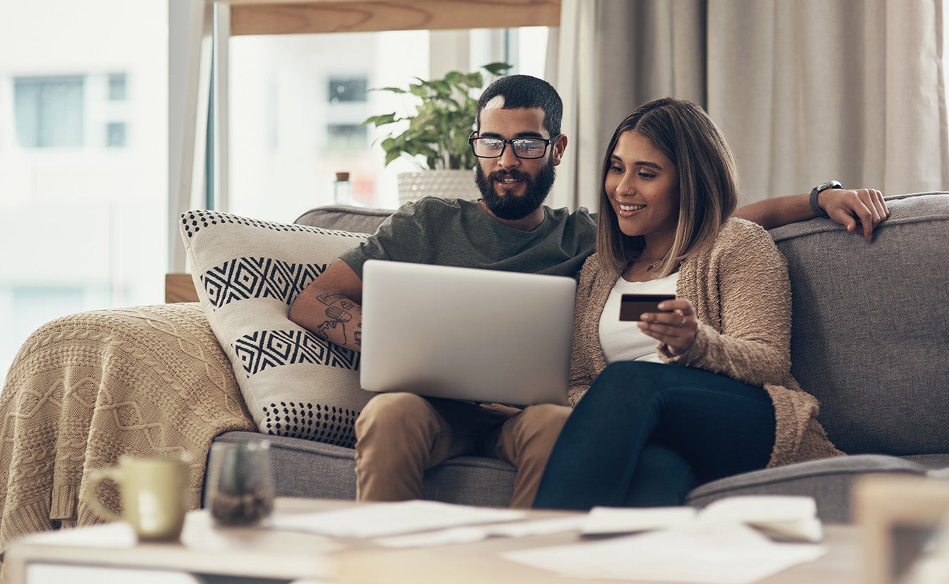 A couple sitting on a couch looking at a laptop, one holding a card for online payment.