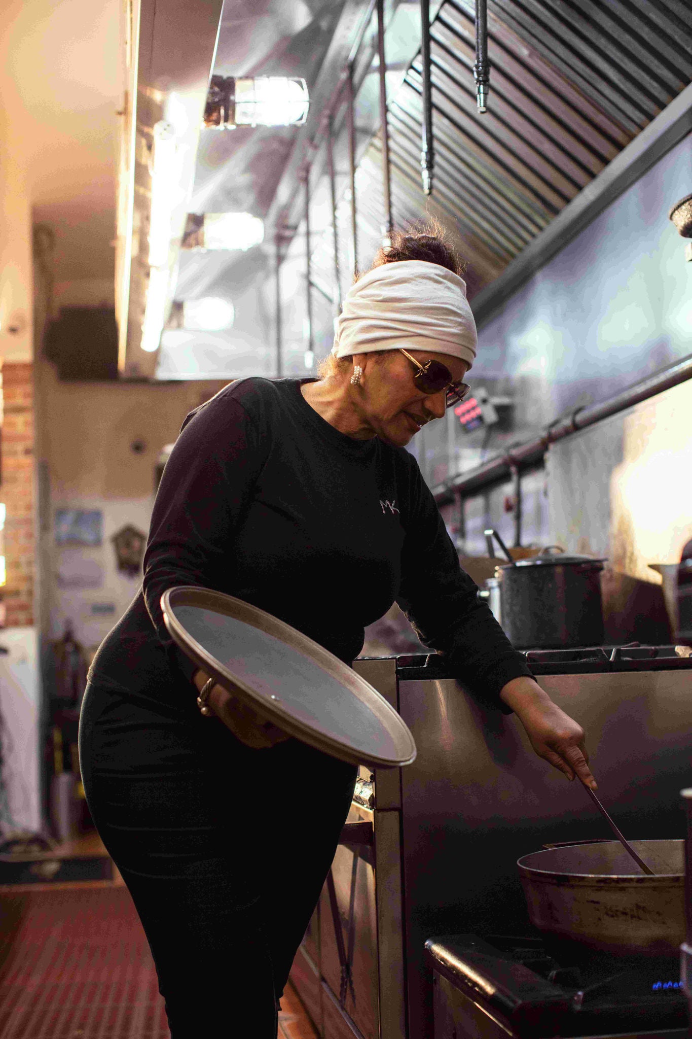  A chef reaches into a pot with a utensil while holding the pot cover in her other hand in a restaurant kitchen. 