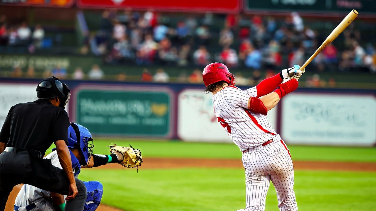 A batter swings his bat in front of a catcher and umpire in a stadium.