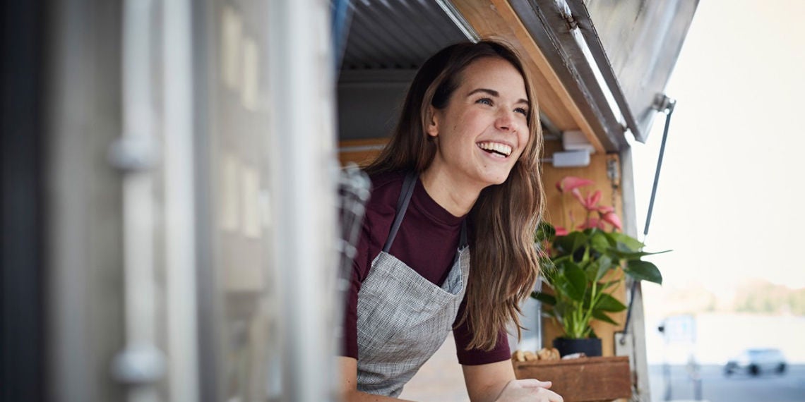 A woman in an apron smiles, leaning out of a window from a food truck.