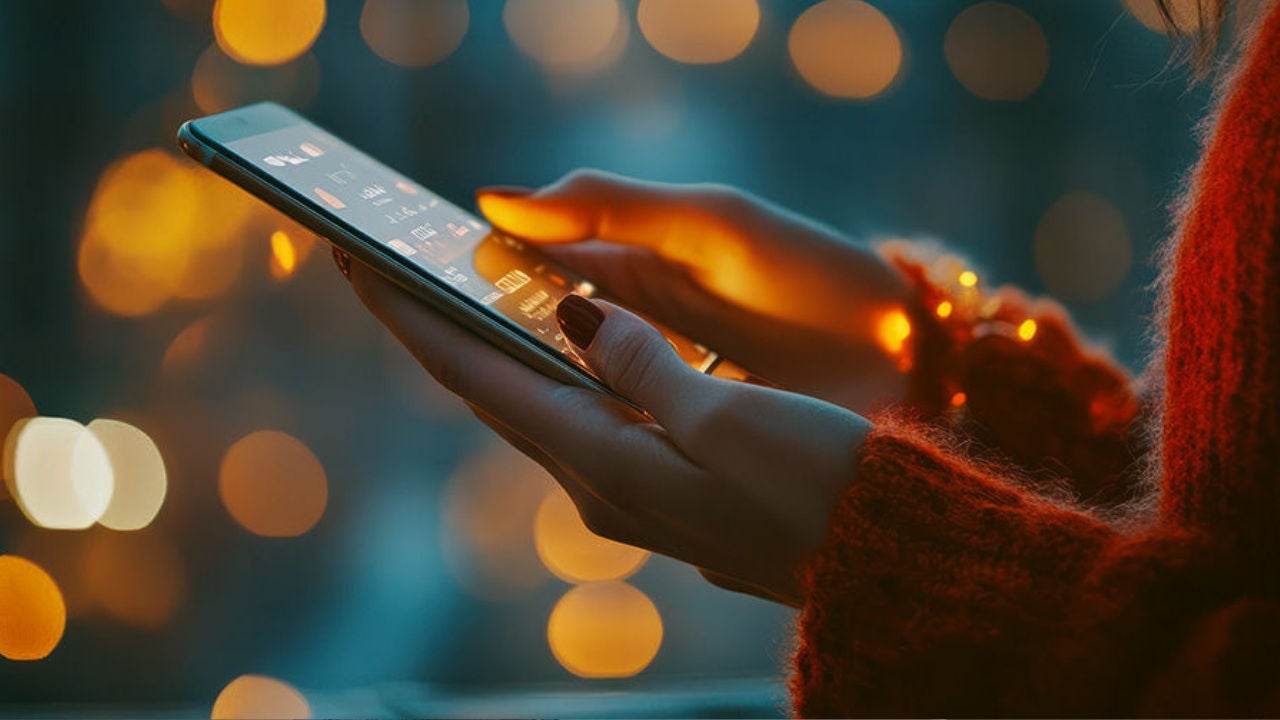 A woman's hand holding the phone against a dark background with multi-colored lights. 