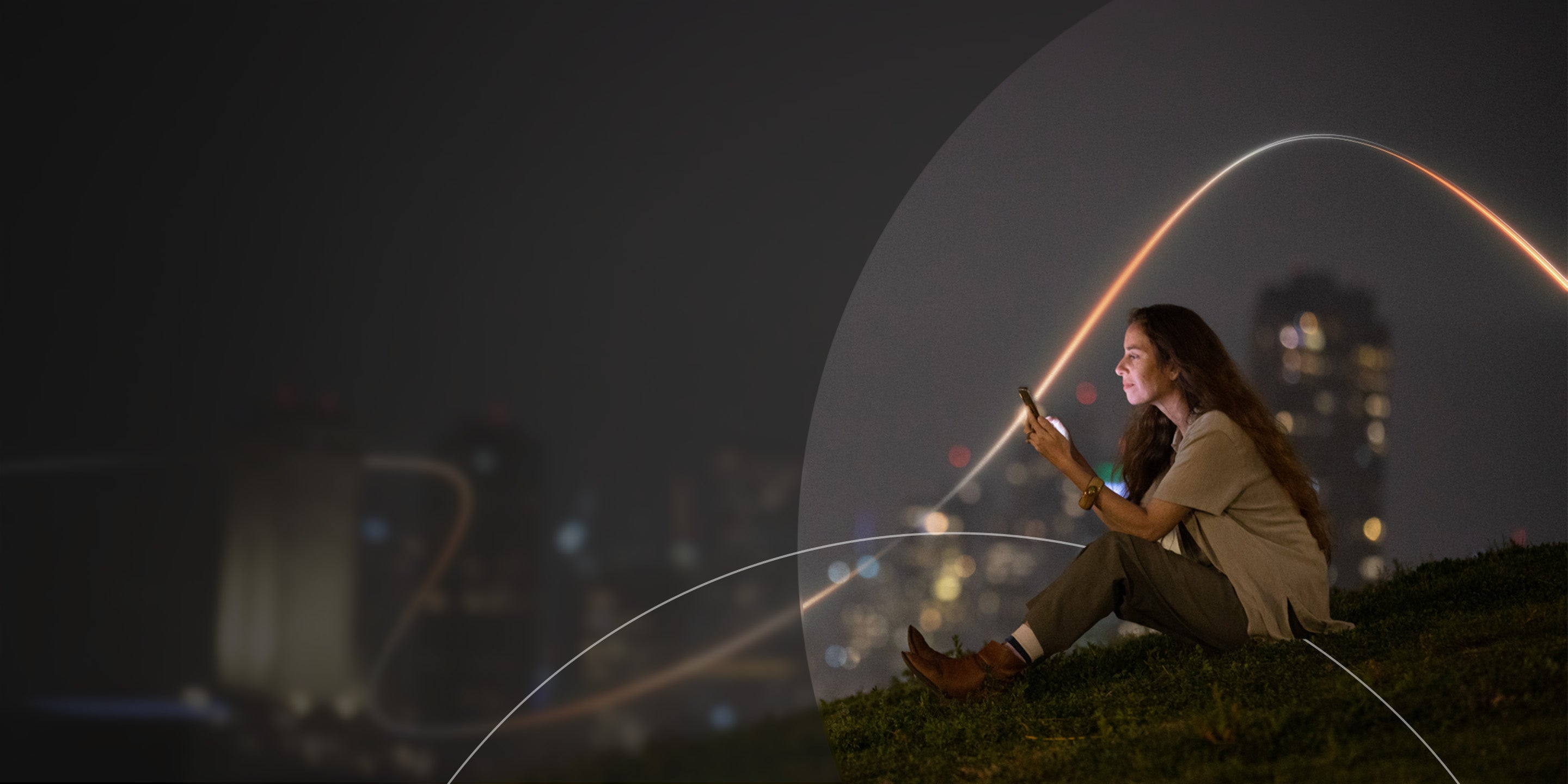 Woman sitting on grass at night, using her phone, with blurred city lights glowing in the background.