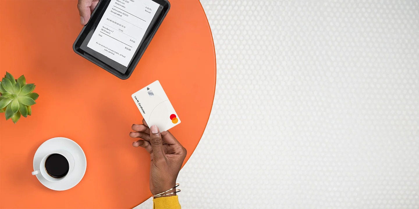 يقوم مستهلك بإجراء عملية شراء باستخدام بطاقة Mastercard للعملات الرقمية، بينما تحمل يد أخرى جهازًا لوحيًا يعرض شاشة الدفع.