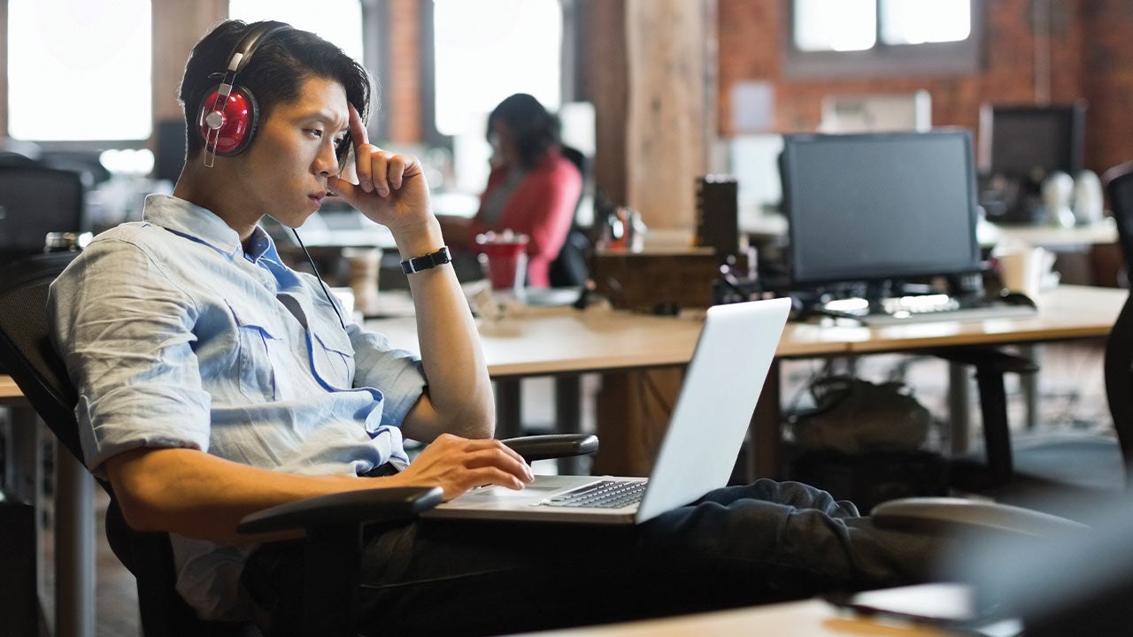 Fejhallgatót viselő férfi nyitott irodában ül, laptopon dolgozik, és koncentráltan a képernyőt figyeli.