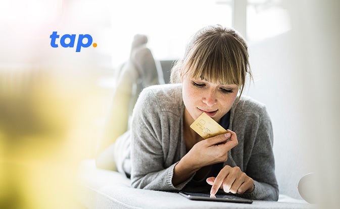 A woman lying on a couch, holding a crypto card in one hand and using the other hand to interact with a tablet device.