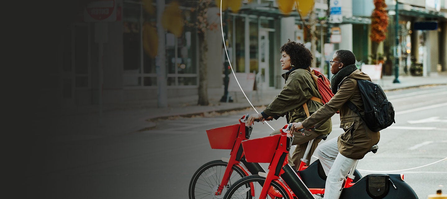 Two people with backpacks riding red bikes on a city street.